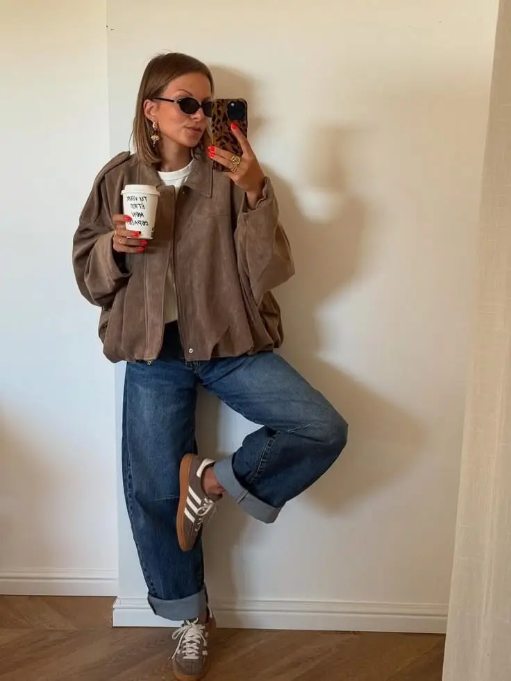 woman mirror selfie in taupe mocha suede blouson jacket white tee rolled medium wash jeans brown Samba style sneakers gold statement earrings oval sunglasses coffee cup outfit 26