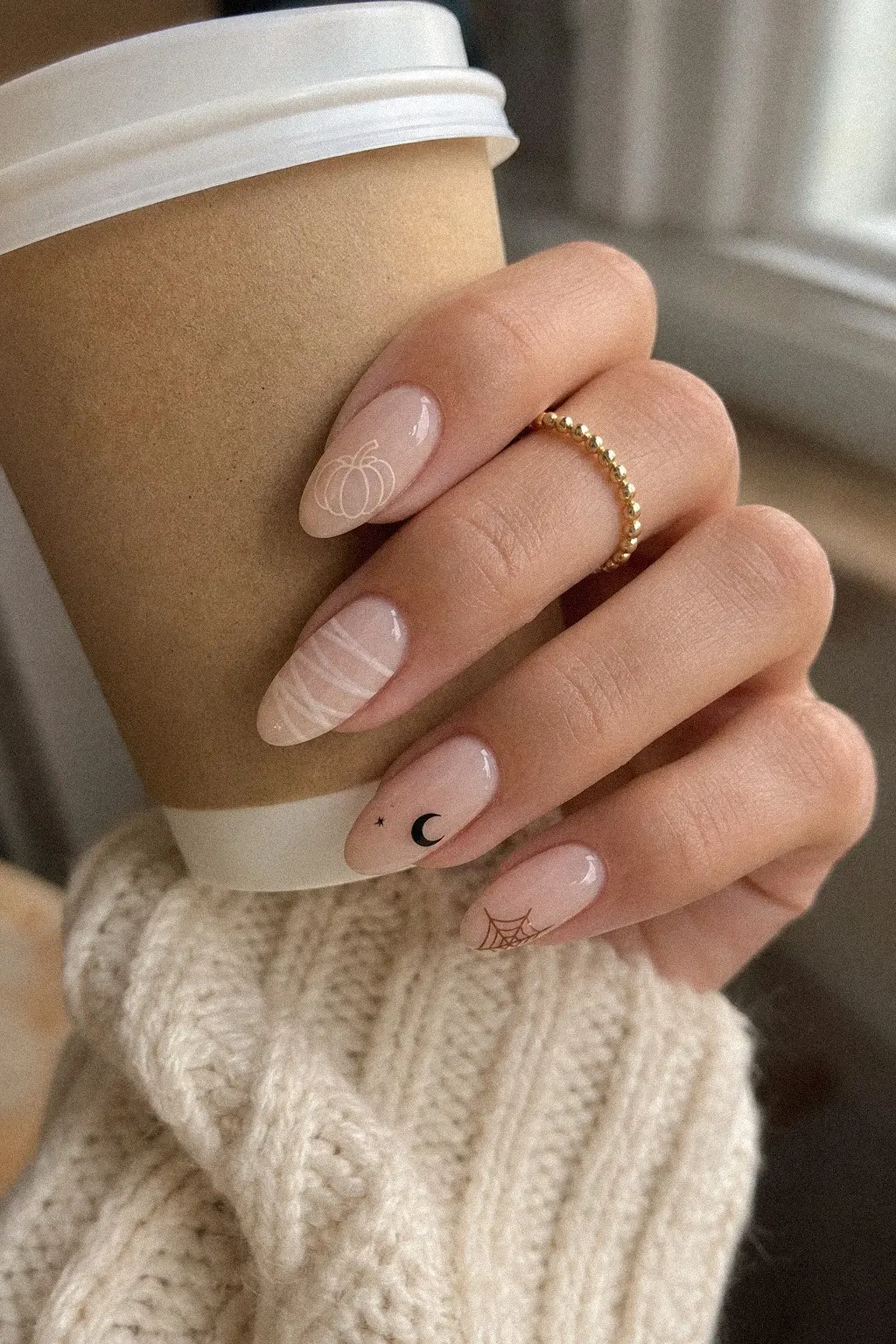 medium almond nails with sheer milky nude gel base, cream pumpkin outline, mummy wrap stripes, crescent moon star, and brown spiderweb corner details, hand on cream knit sweater, look 1