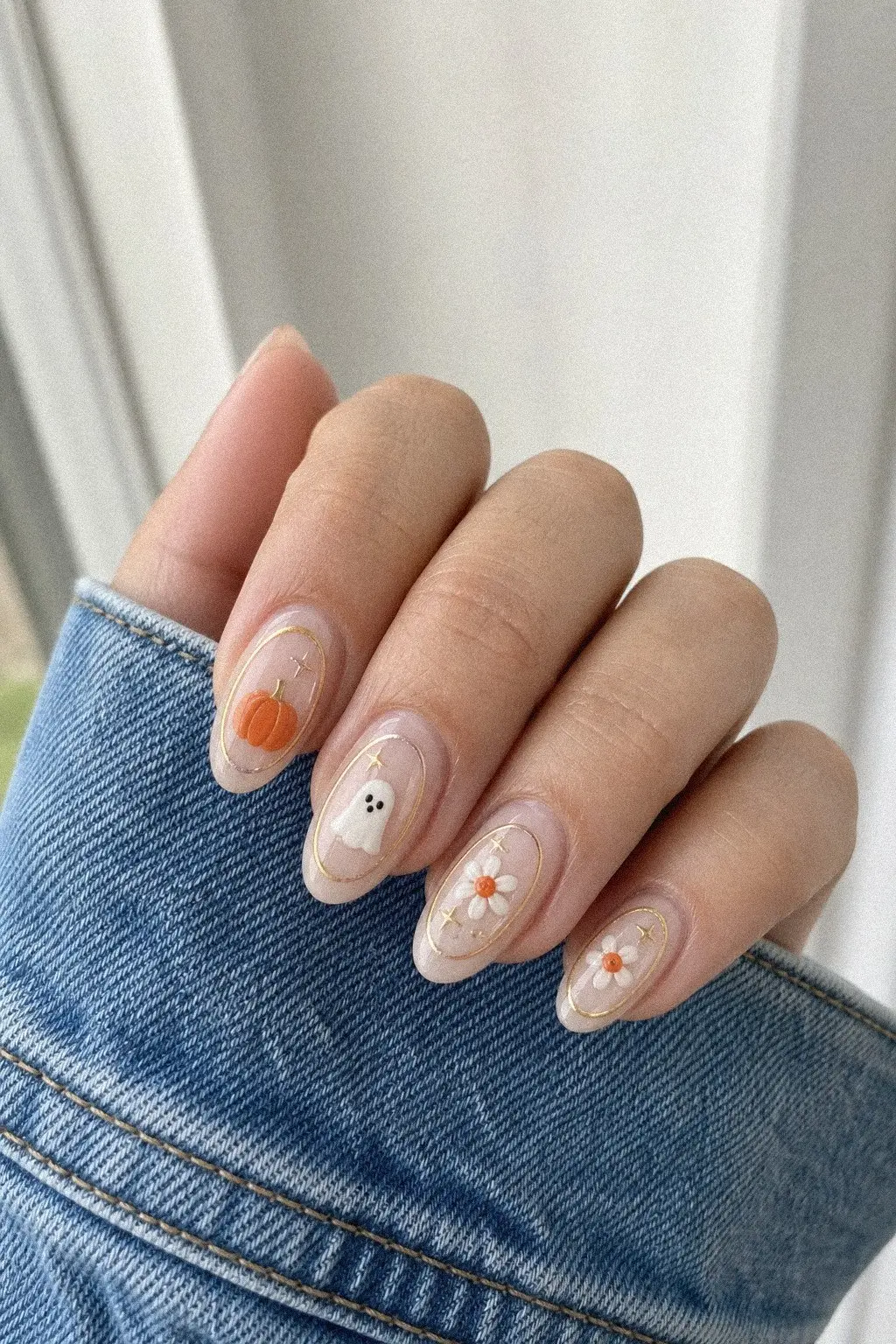 medium almond nails with milky pink gel base, thin gold oval frame outlines, 3D style orange pumpkin, white ghost, white daisies with orange centers details, hand on white ribbed fabric, look 2