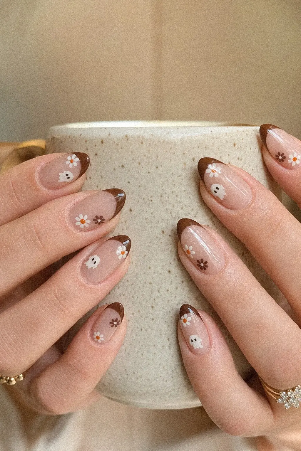 medium almond nails with sheer nude base, rich chocolate brown French tips, tiny white ghost nail art, white and orange daisy details, hands with gold and diamond rings, look 3