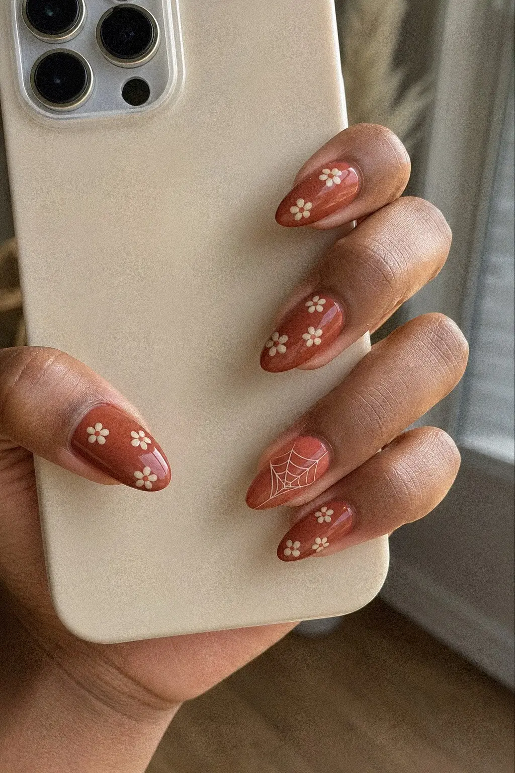 medium almond nails with opaque terracotta gel base, cream daisy florals and thin spiderweb corner detail, hand holding phone nails facing camera, look 5