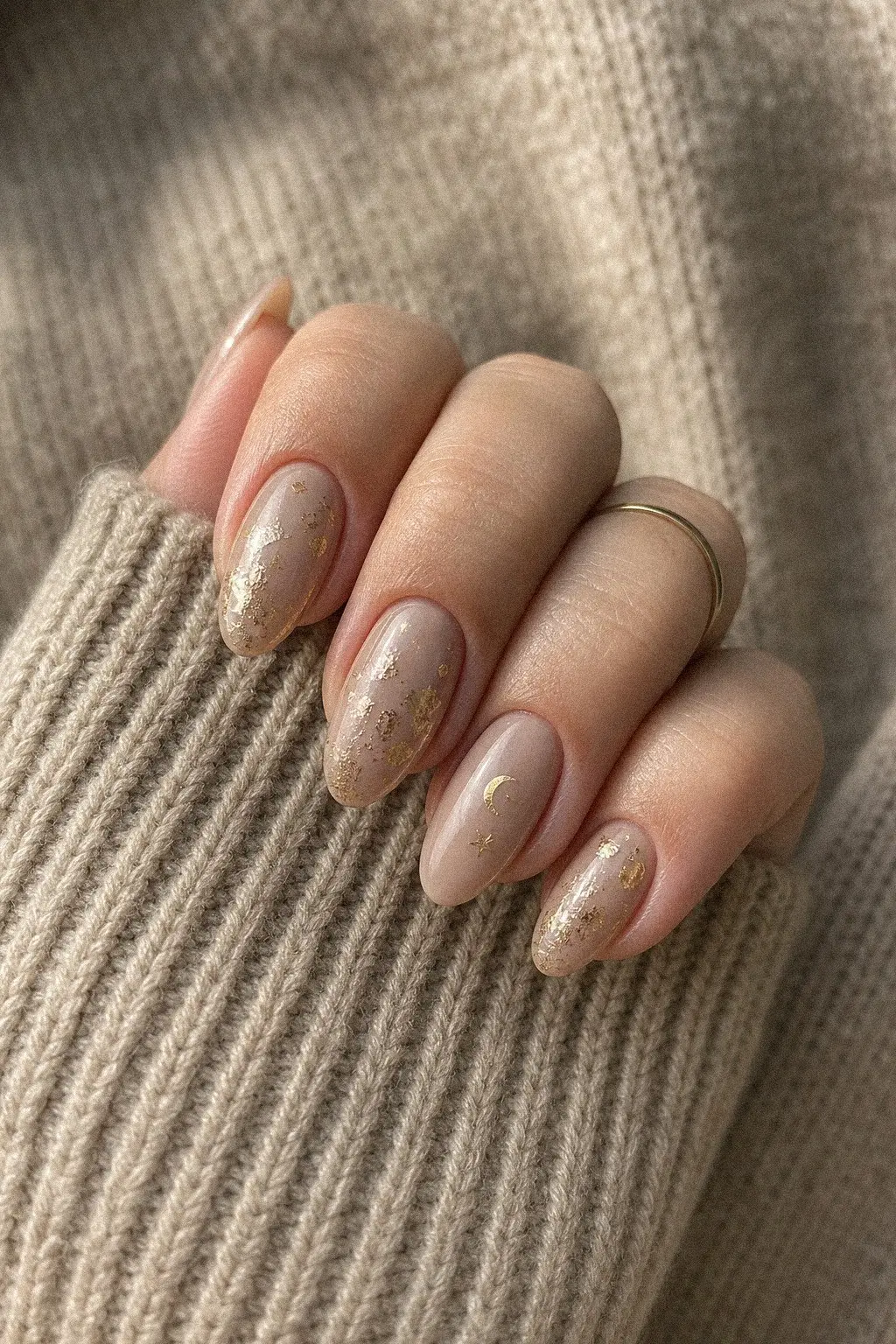 medium almond nails with warm greige nude gel base, scattered gold foil leaf shapes, crescent moon and star accent on one nail in gold, hand on knit sweater sleeve, look 6
