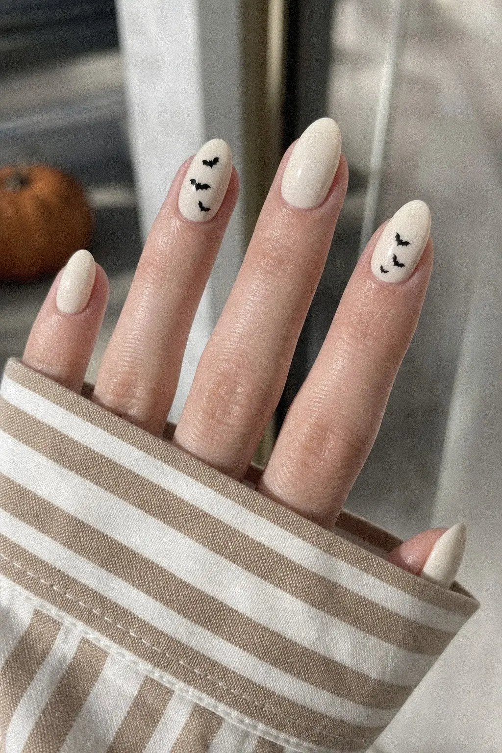 medium almond nails with warm cream off-white gel base, small black bat silhouettes on accent nails, fingers spread over striped shirt cuff, look 7