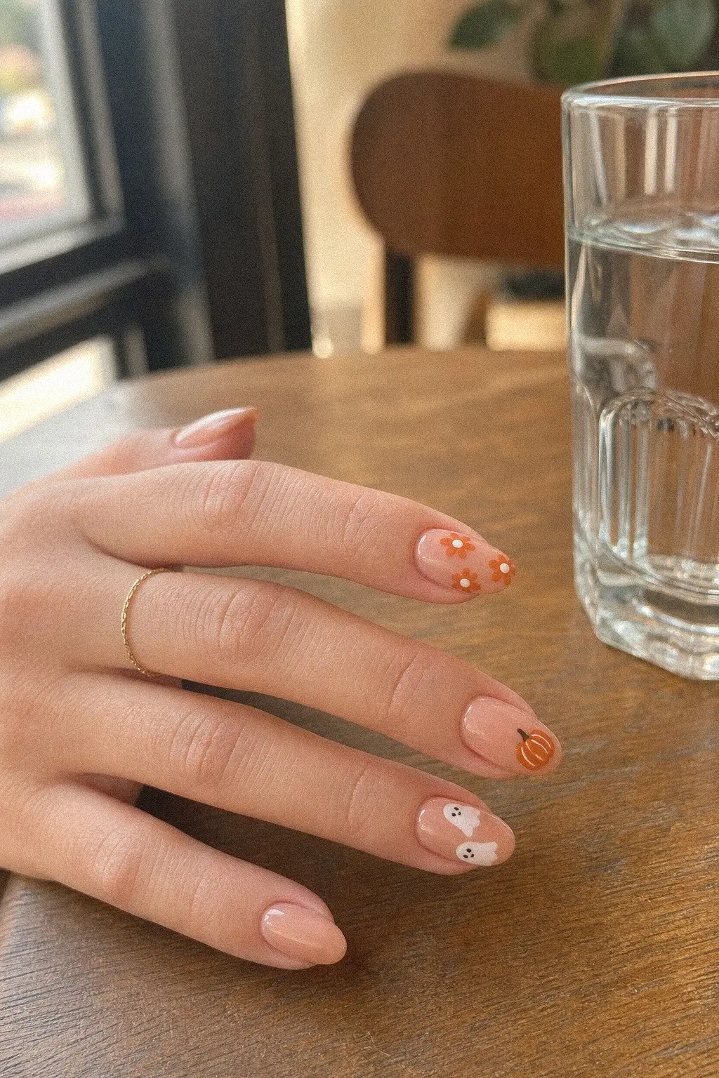 medium almond nails with warm blush peach gel base, burnt orange daisy florals, orange pumpkin accent, white ghost pair with black eyes, hand resting on cafe table, look 8