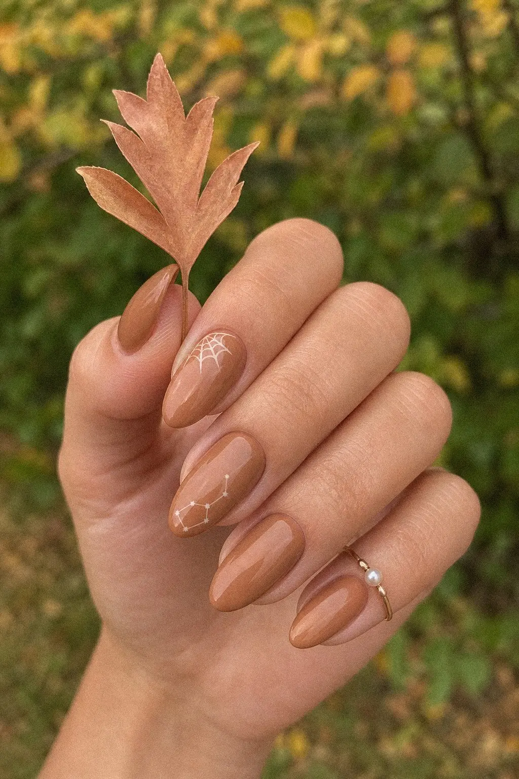 medium almond nails with opaque caramel latte gel base, cream spiderweb corner accent, tiny constellation star map on accent nail, hand holding a small leaf, look 9