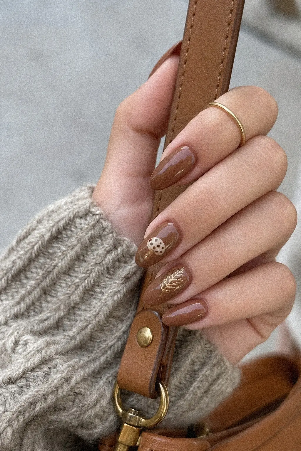 medium almond nails with warm brown gel base, cream and brown mushroom motif accent nail, golden autumn leaf outline detail, hand on leather handbag strap with rings, look 10