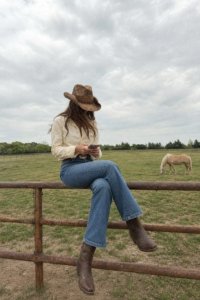 woman in cream linen button shirt and high-rise flare jeans with silver concho belt and brown woven cowboy hat sitting on a ranch fence with horses in the background, outfit 2