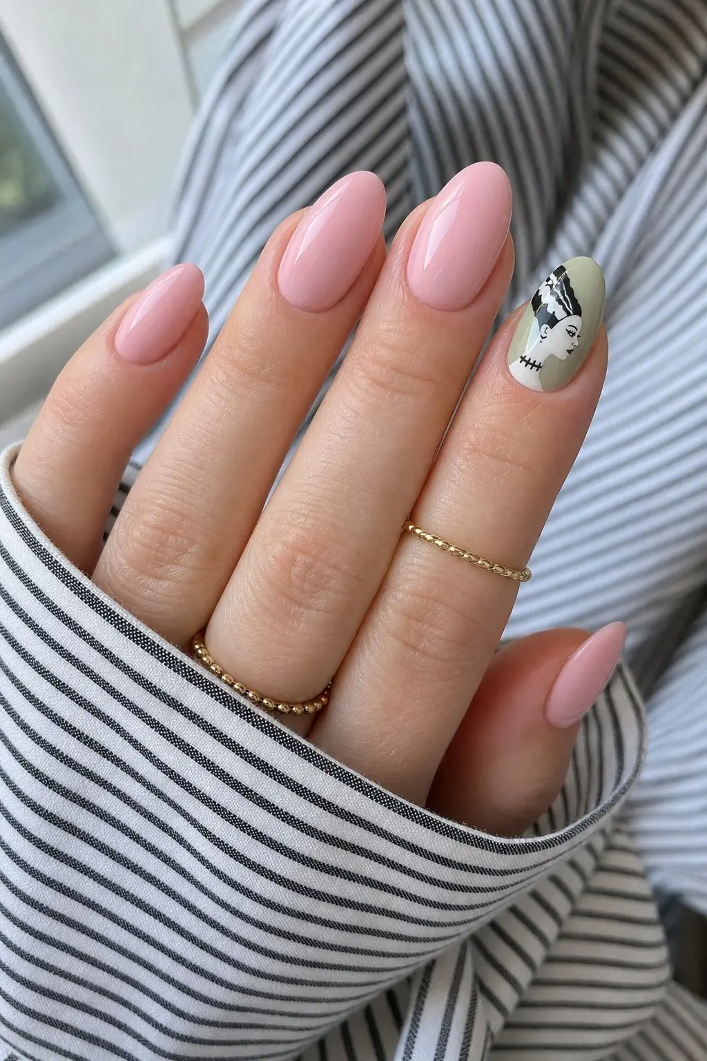 medium almond nails with blush pink gel on most fingers, pale sage green accent nail with Bride of Frankenstein face painted in black and white, gold chain rings on fingers, nail look 17