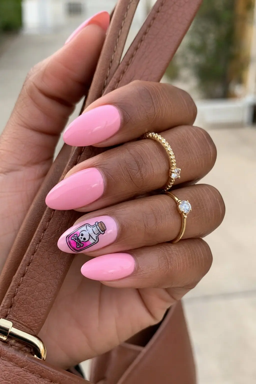 medium almond nails with bubblegum pink gel base on most fingers, white accent nail on ring finger with hand-painted pink poison bottle and skull crossbones label, gold stacked rings, nail look 20
