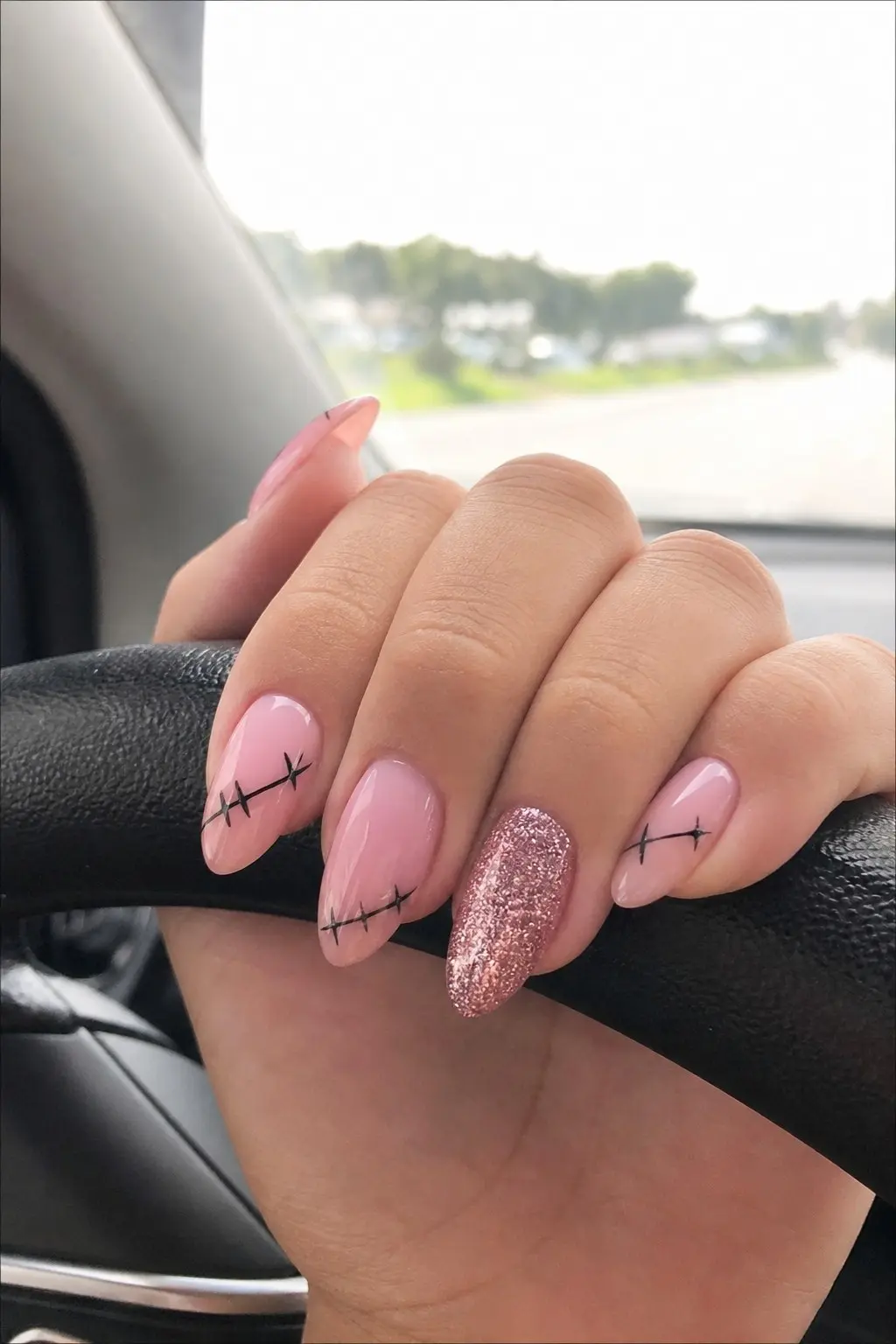 medium almond nails with soft pink gel base and black barbed wire stitch line design across most nails, one ring finger accent nail in metallic pink glitter gel, nail look 24