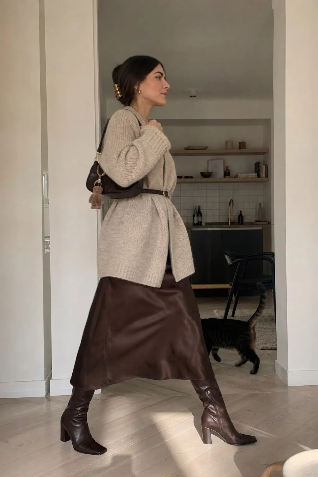 woman in oatmeal chunky ribbed oversized sweater belted dark brown leather belt chocolate faux leather midi skirt dark brown knee high heeled boots dark brown shoulder bag with charm gold hair claw clip outfit 10
