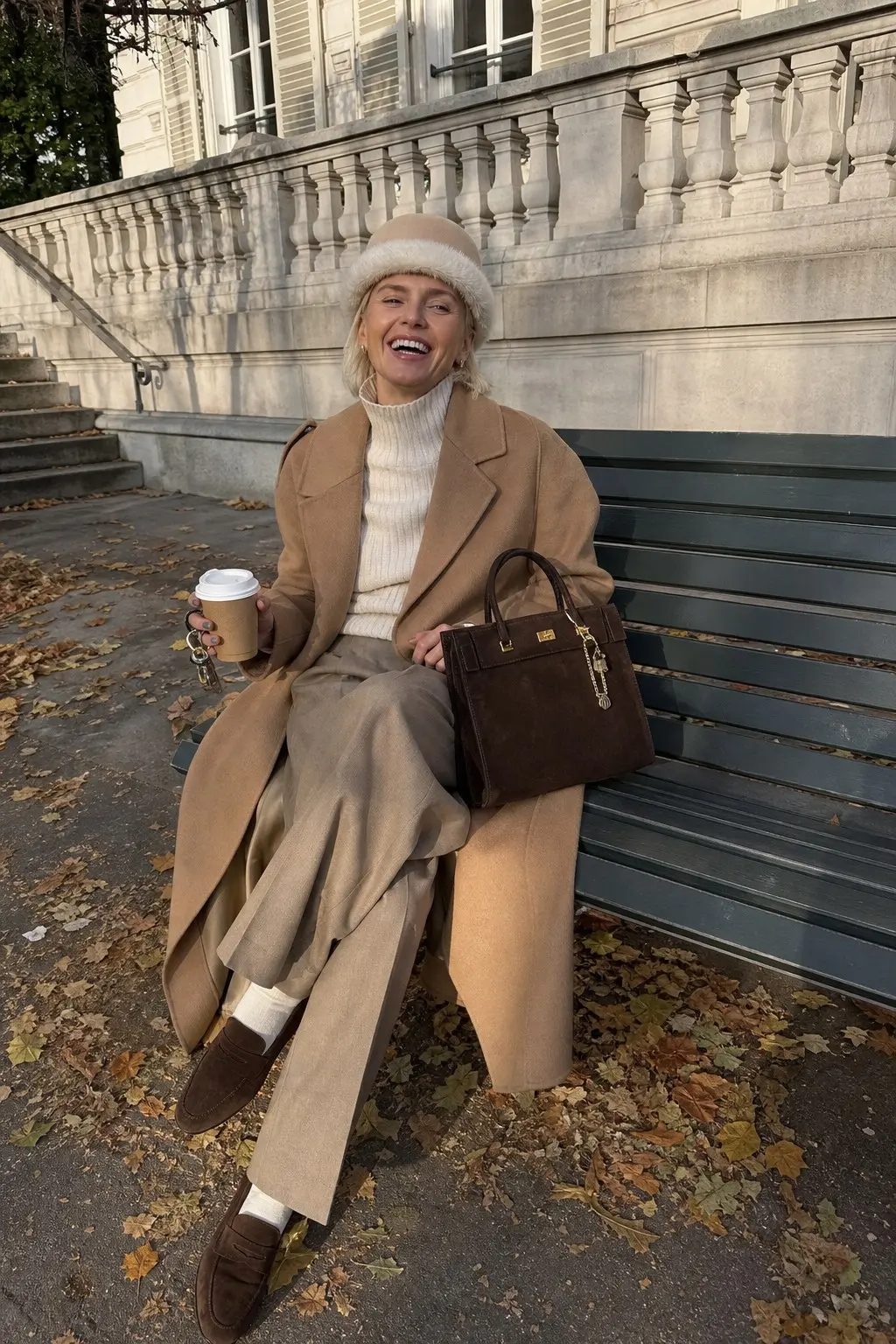 woman in long camel wool coat cream ribbed turtleneck taupe wide leg trousers dark brown suede loafers white crew socks fuzzy beige beret dark brown suede structured bag outfit 21