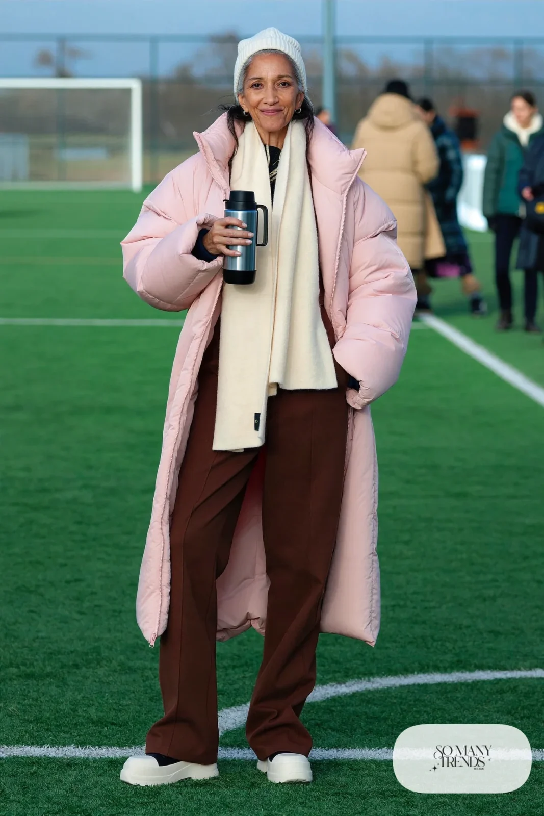 Woman in her fifties in a long pale pink puffer coat, espresso ponte pants, cream knit hat and white boots, outfit 12