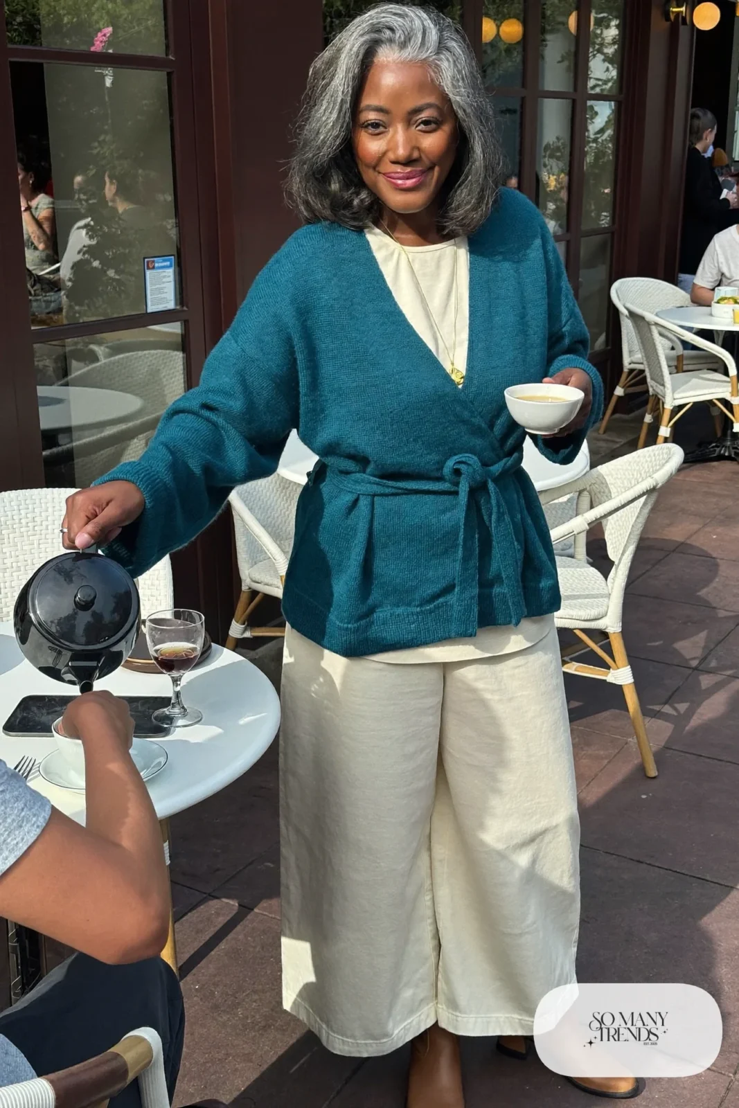 Woman in her fifties in a teal wrap cardigan, cream tee, wide cream pants and brown ankle boots, outfit 16