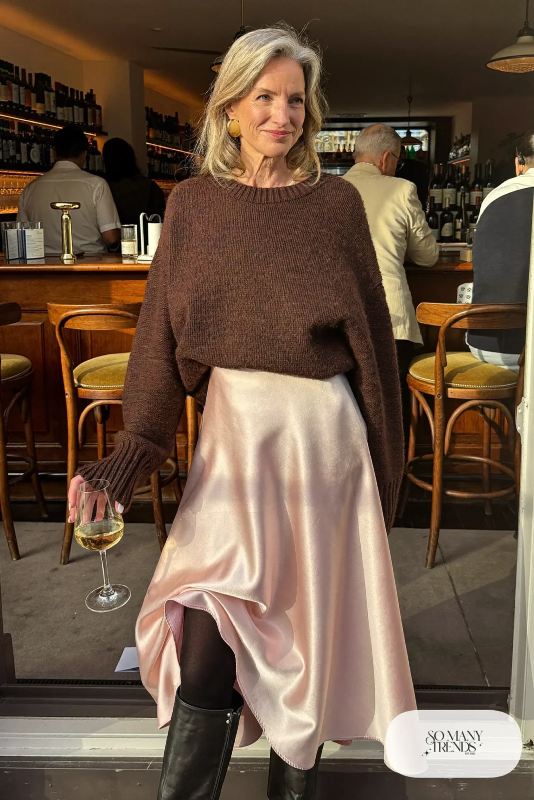 Woman in her fifties in a pale pink satin midi skirt, chunky espresso sweater, tights and tall boots, outfit 17