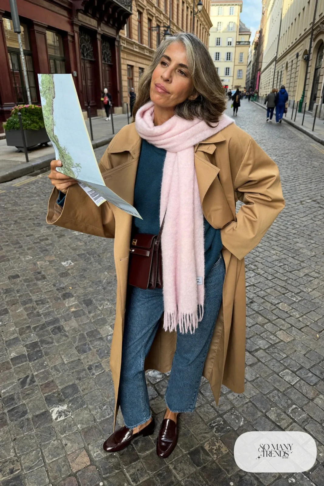 Woman in her fifties in a camel trench coat, teal sweater, straight jeans and loafers on a city street, outfit 31