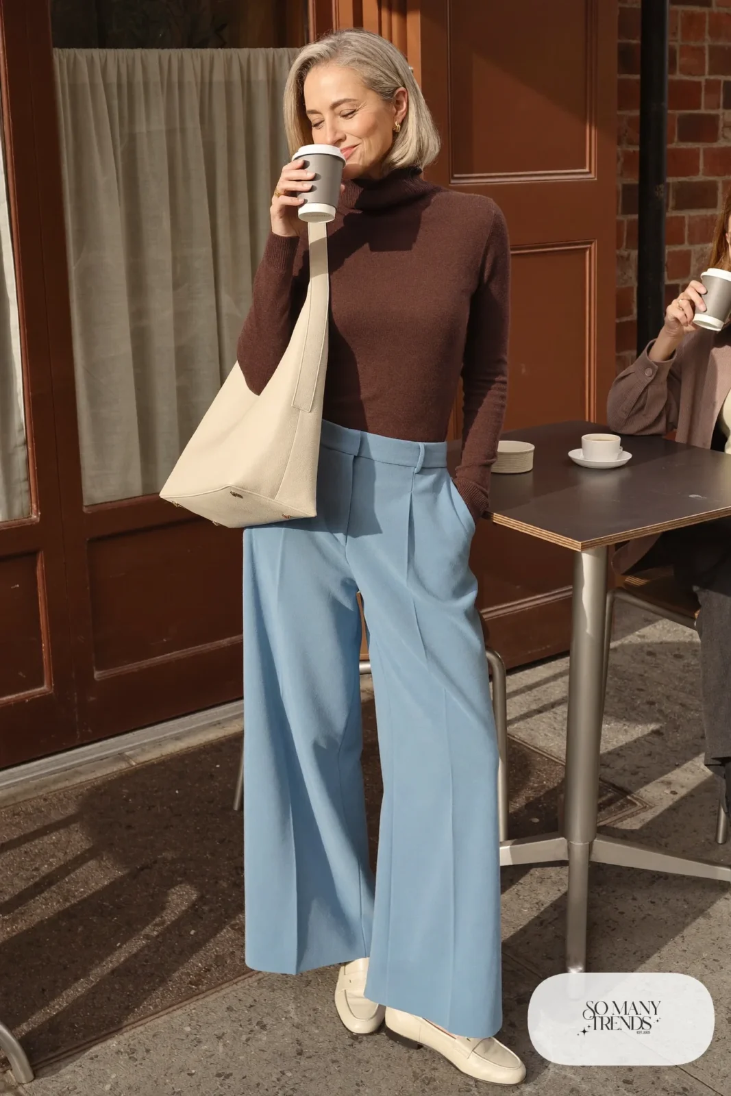 Woman in her sixties wearing a chocolate brown ribbed turtleneck, powder blue wide-leg pants, cream loafers and a cream leather bag, outfit 1