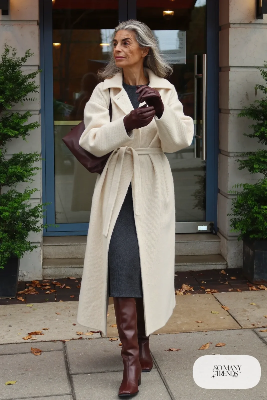 Woman in her sixties wearing a cream belted wrap coat over a charcoal knit midi dress and brown tall boots, outfit 7