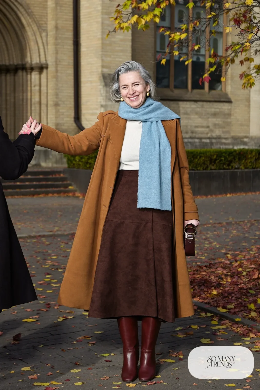 Woman in her sixties wearing a chocolate suede midi skirt, a cream turtleneck, a powder blue scarf and tall brown boots, outfit 12