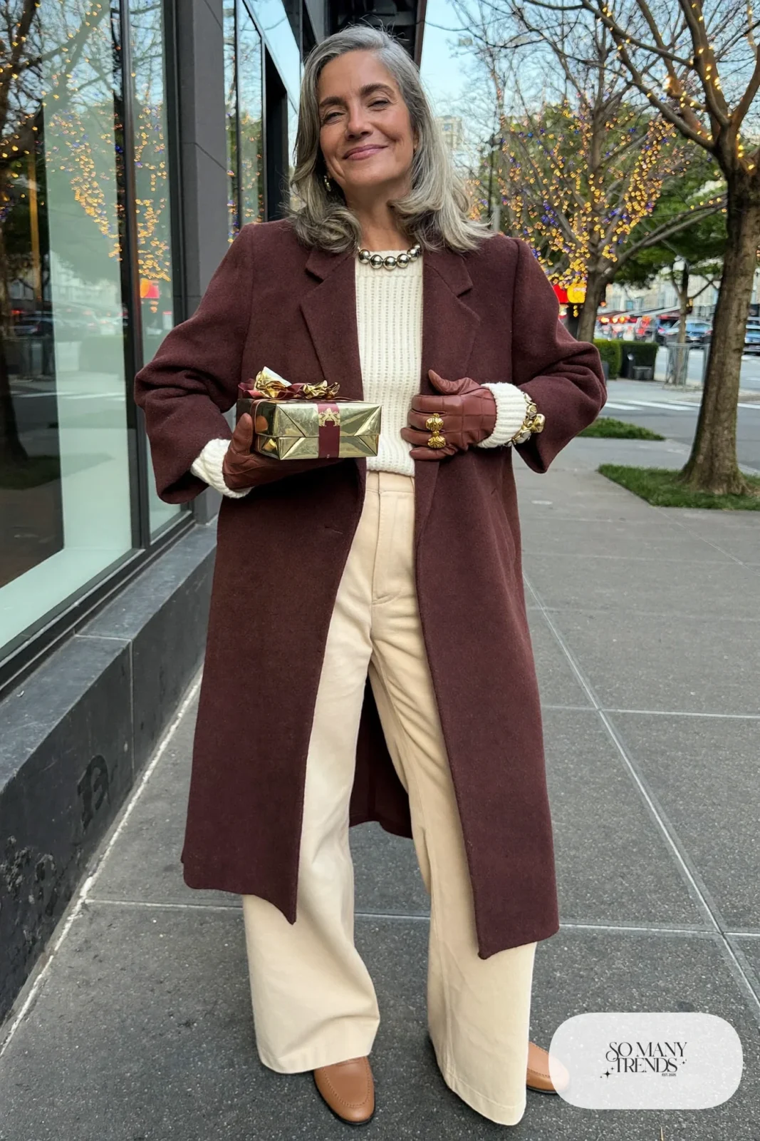 Woman in her sixties wearing a burgundy long wool coat over a cream sweater and cream wide-leg pants with brown gloves, outfit 18