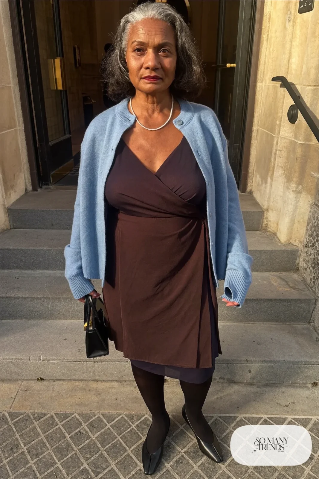Woman in her sixties wearing a navy wrap midi dress, a powder blue cardigan, sheer tights and black low pumps, outfit 19