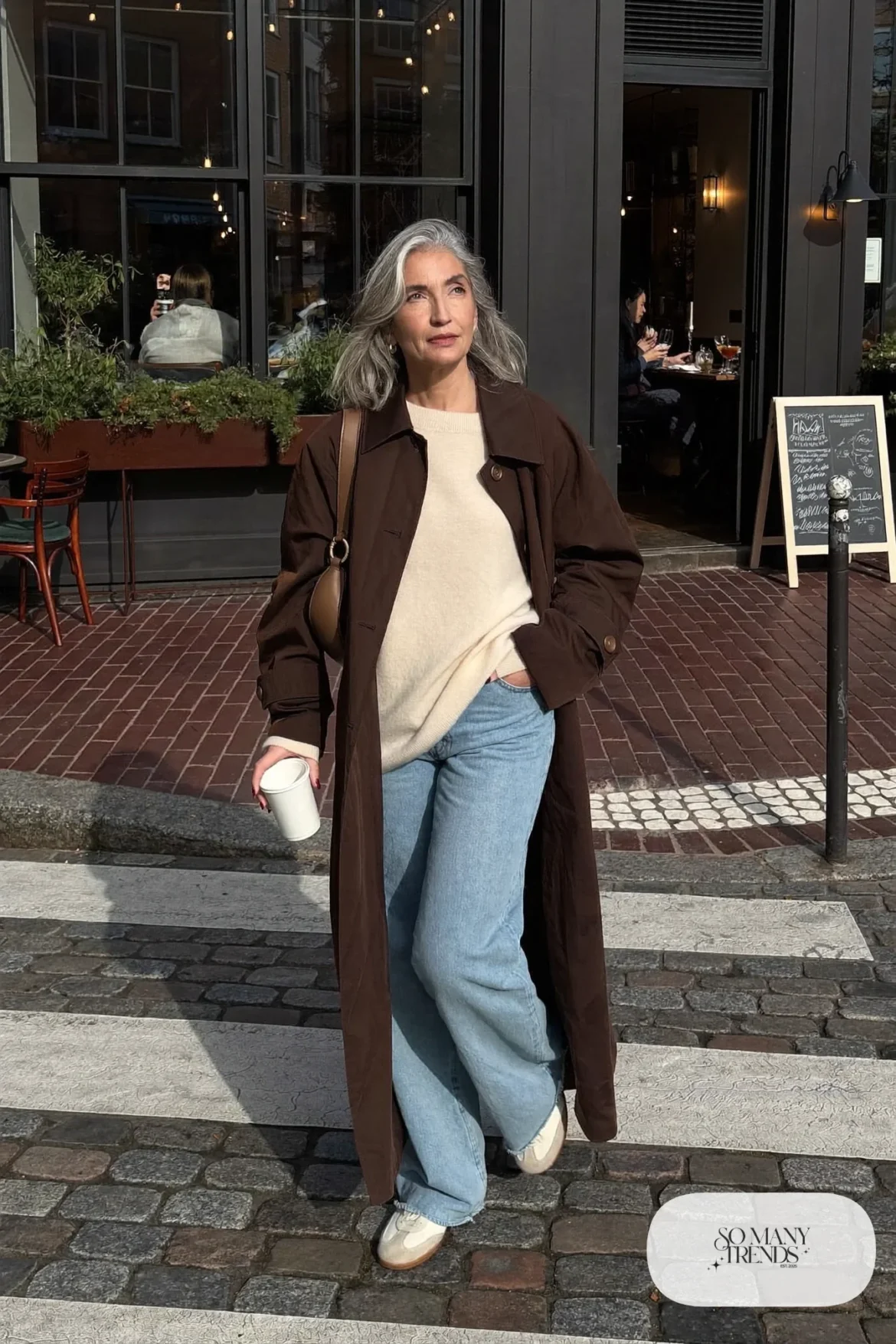 Woman in her sixties wearing a chocolate brown trench coat, a cream knit, light wide-leg jeans and cream sneakers, outfit 20