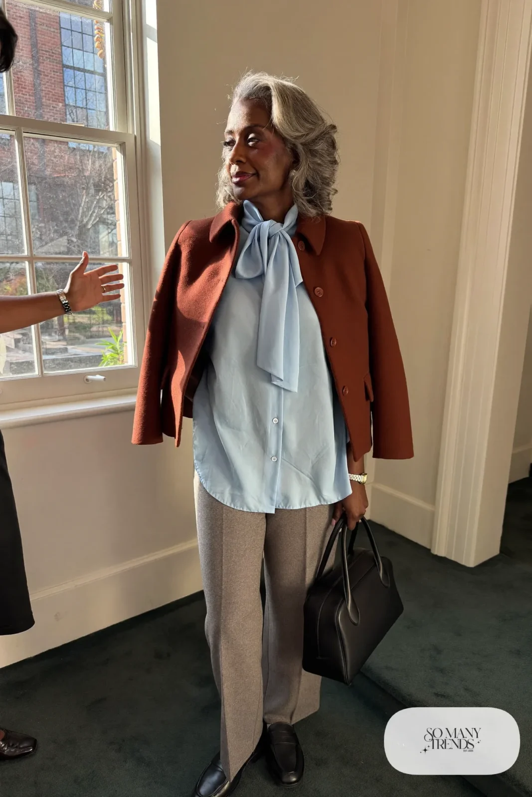 Woman in her sixties wearing gray flannel pants, a powder blue blouse with a soft bow and black loafers, outfit 23