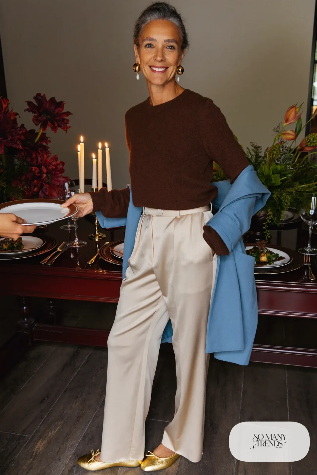 Woman in her sixties wearing cream satin wide-leg pants, a chocolate knit sweater, gold flats and drop earrings, outfit 25
