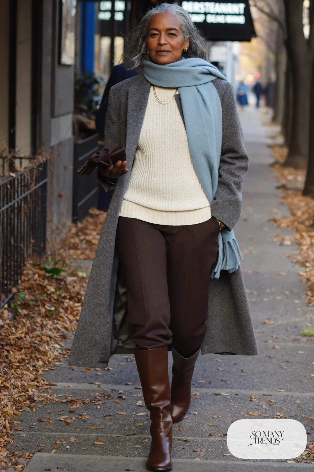 Woman in her sixties wearing a long gray coat, a powder blue scarf, a cream sweater, chocolate pants and tall brown boots, outfit 29