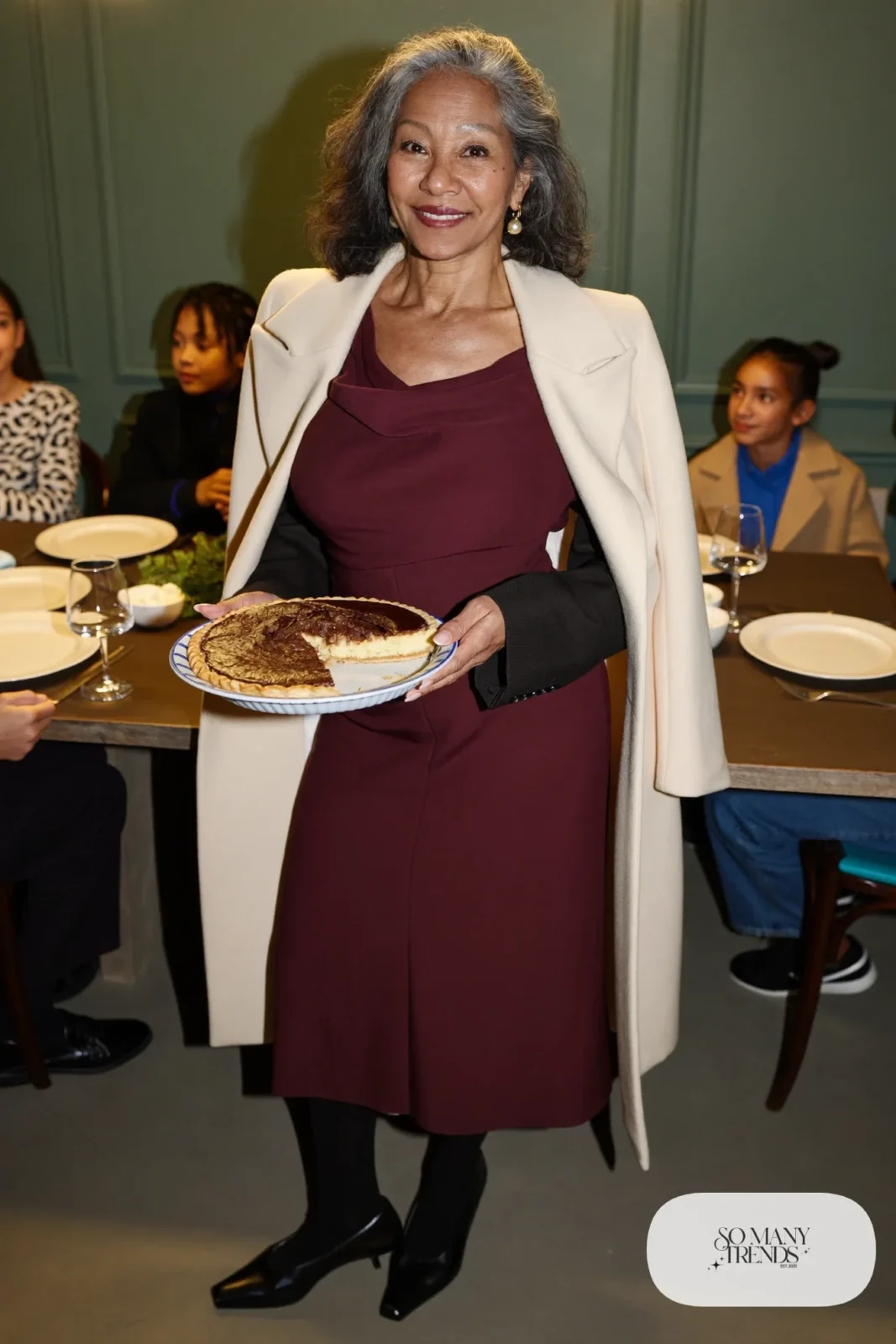 Woman in her sixties wearing a burgundy jersey midi dress with a draped neckline, sheer tights and low black pumps, outfit 30
