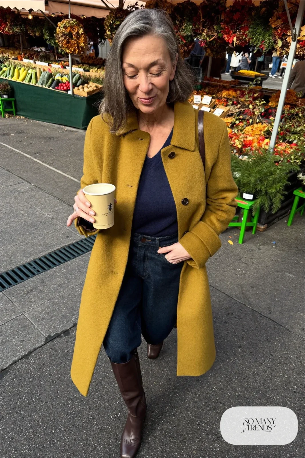 Petite woman in a belted mustard knee-length coat over a navy sweater and dark jeans with brown boots, outfit 15