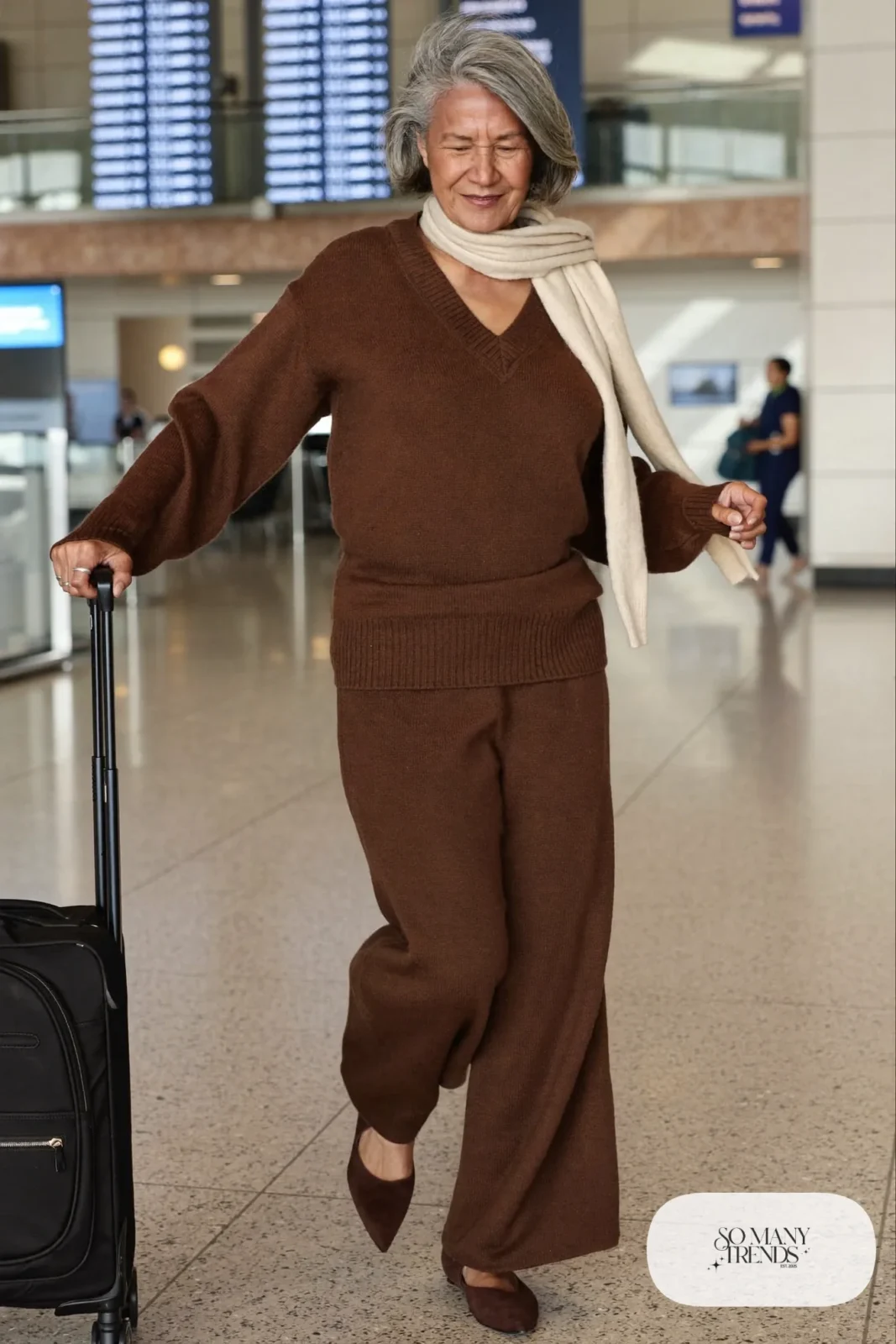 Petite woman in a brown knit V-neck sweater and matching wide-leg knit pants with pointed slip-on flats at an airport, outfit 19