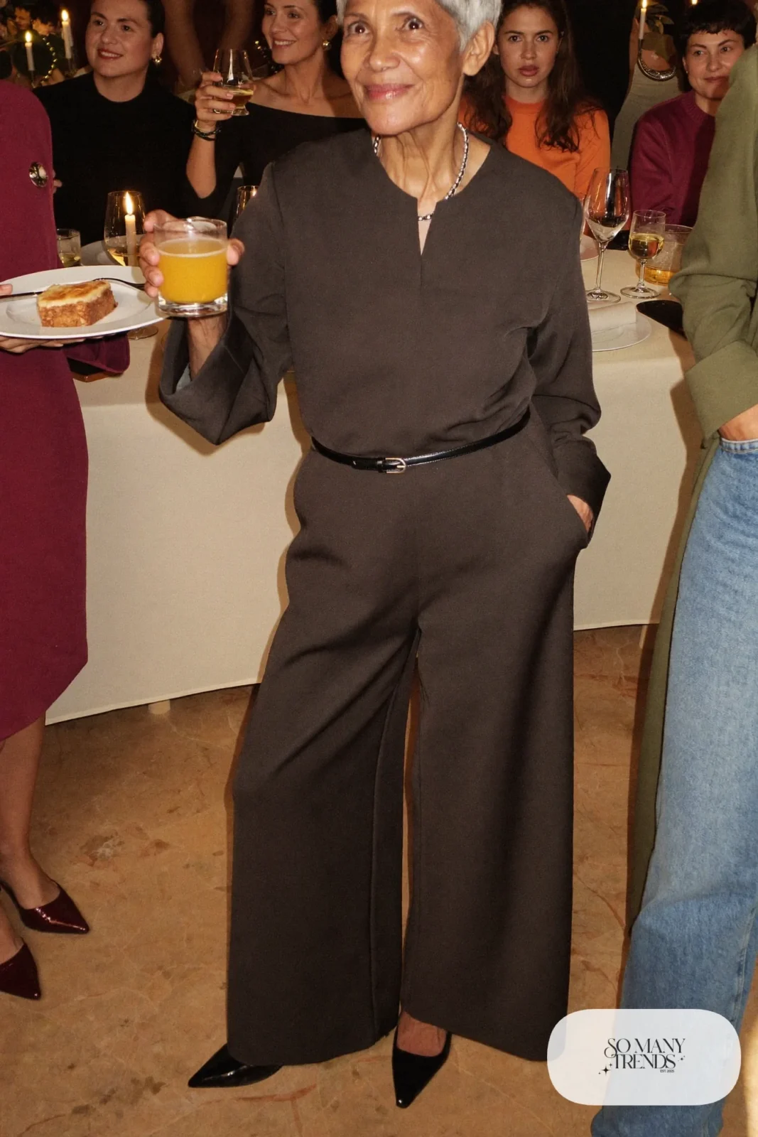 Petite woman in a charcoal wide-leg V-neck jumpsuit with a thin belt, silver necklace and black pumps at a New Year dinner, outfit 30