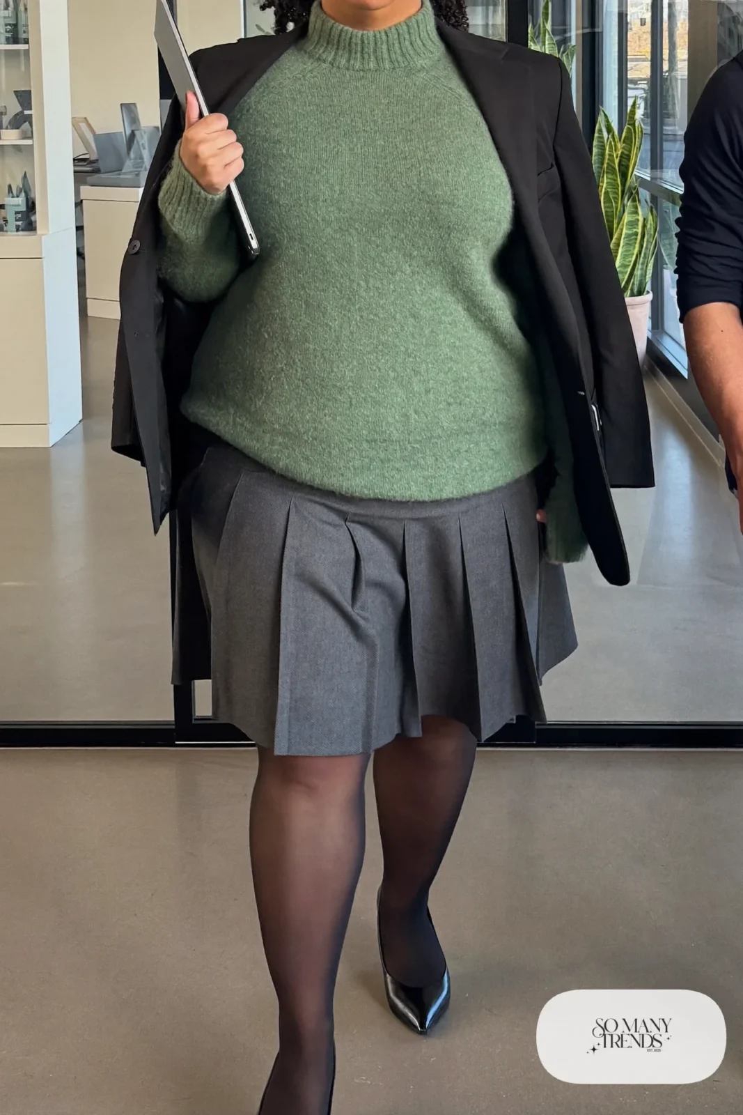 Woman in sage mock-neck sweater, charcoal pleated midi skirt, black blazer and pointed flats, outfit 5