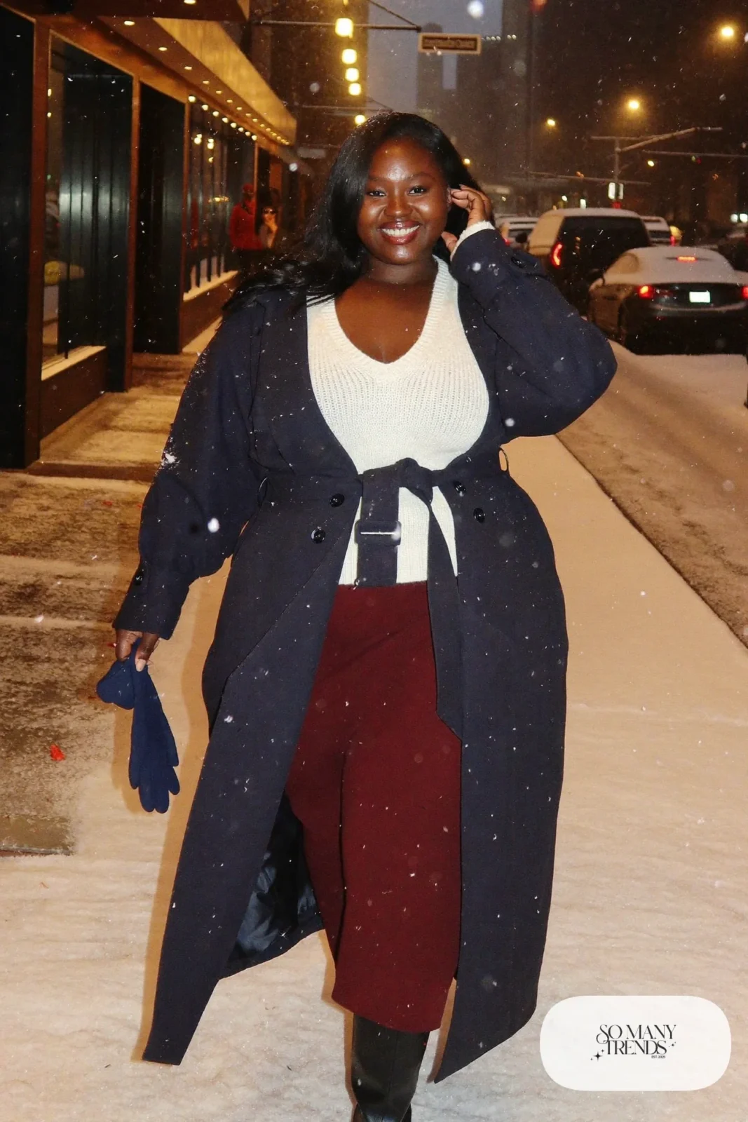 Plus size woman in belted navy long coat, ivory sweater, oxblood skirt and black boots on snowy street, outfit 30
