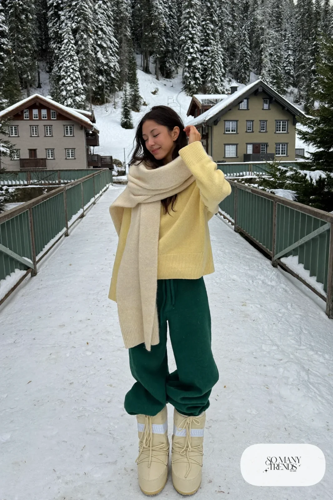 Woman in butter yellow ribbed knit sweater, forest green wide-leg pants, cream moon boots and a cream scarf in the snow, outfit 5