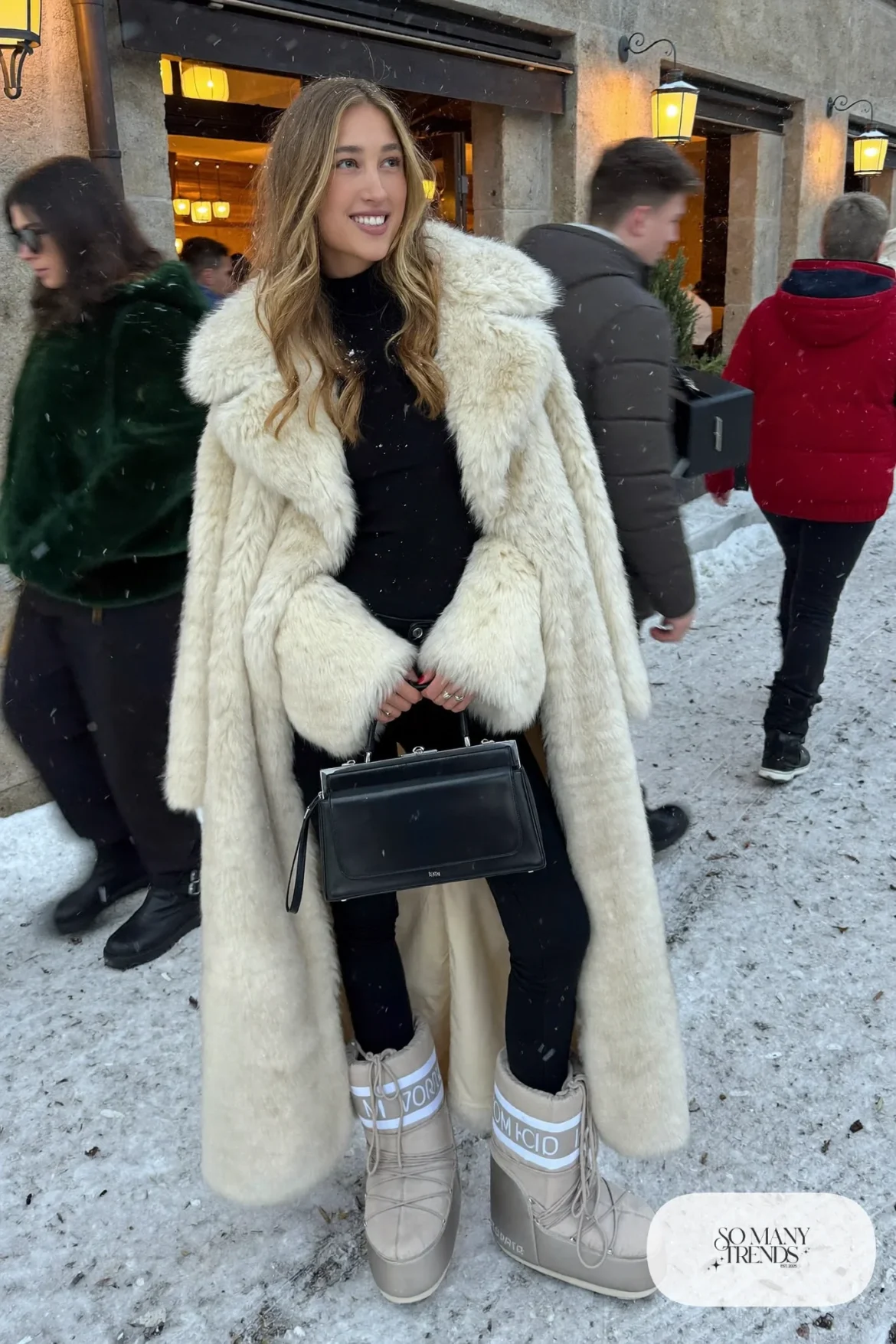 Woman in long cream faux fur coat, black slim jeans, silver moon boots and a black turtleneck in a snowy street, outfit 6
