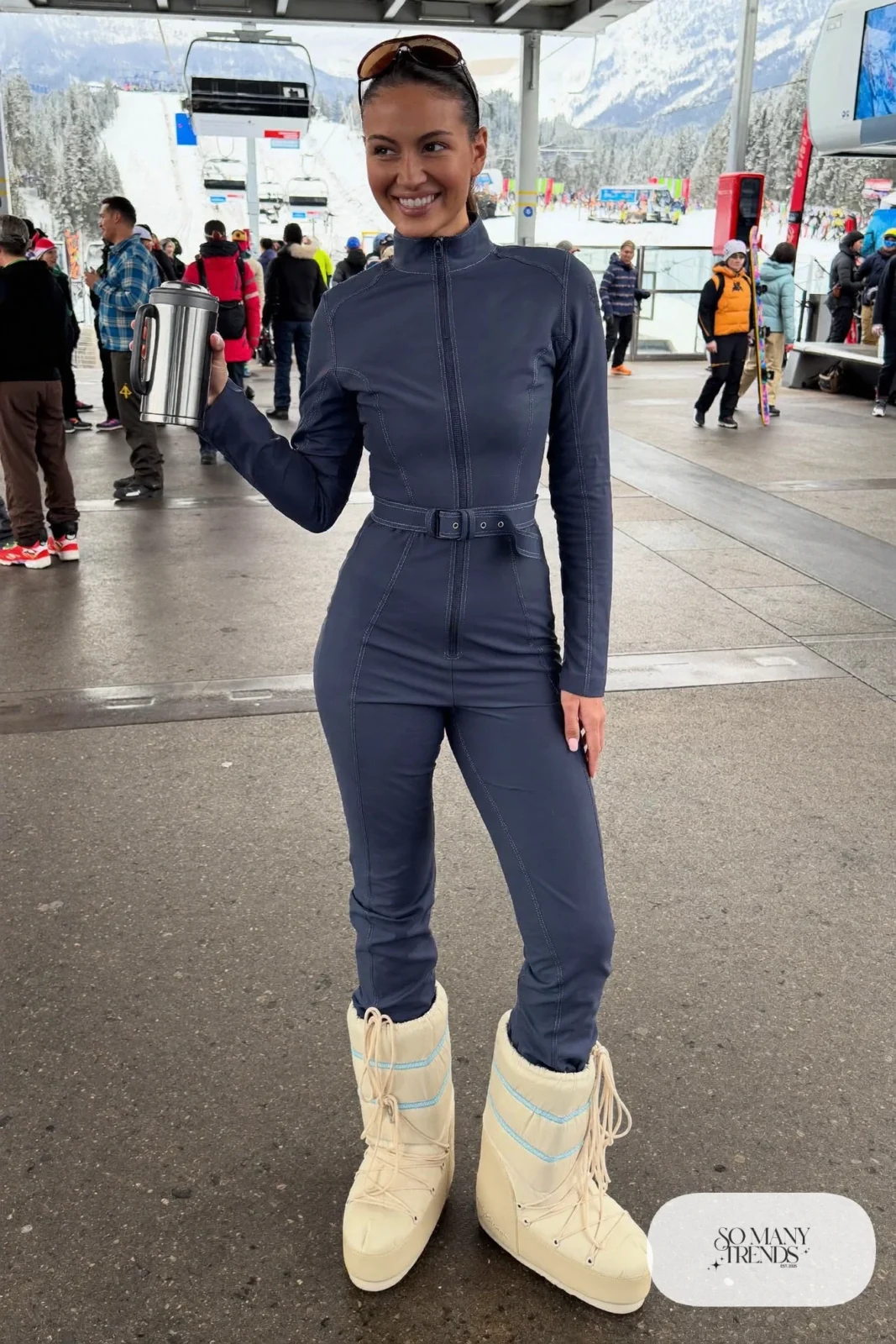 Woman in indigo ski jumpsuit with belt, cream moon boots and a terracotta beanie near a gondola, outfit 8