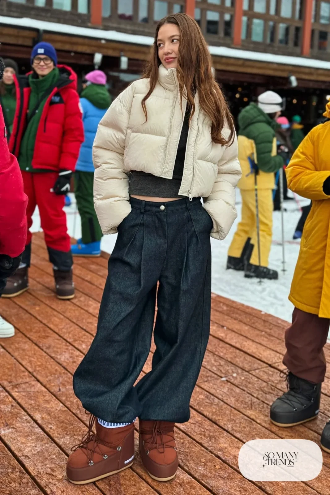 Woman in cropped cream puffer jacket, high-waist indigo wide pants, fitted knit top and brown moon boots in the snow, outfit 11