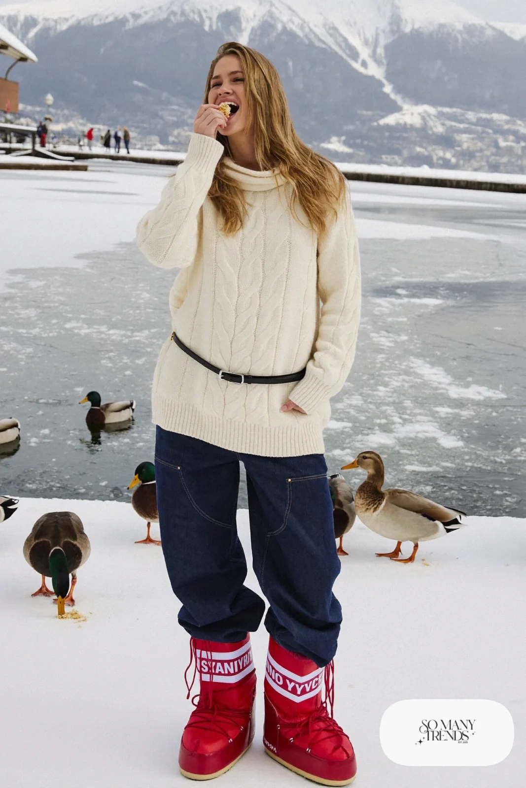 Woman in cream fisherman sweater, barrel-leg indigo jeans, cherry red moon boots and a black belt in the snow, outfit 21