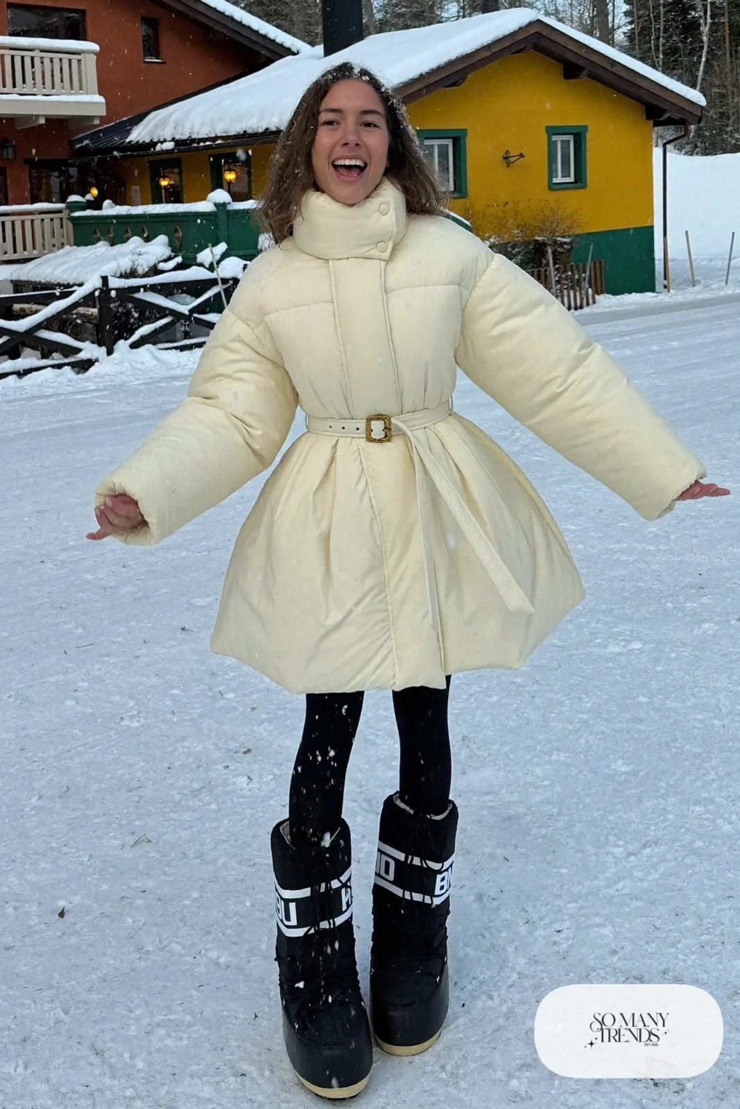 Woman in long cream puffer dress, black tights, black moon boots and a terracotta beanie in the snow, outfit 24
