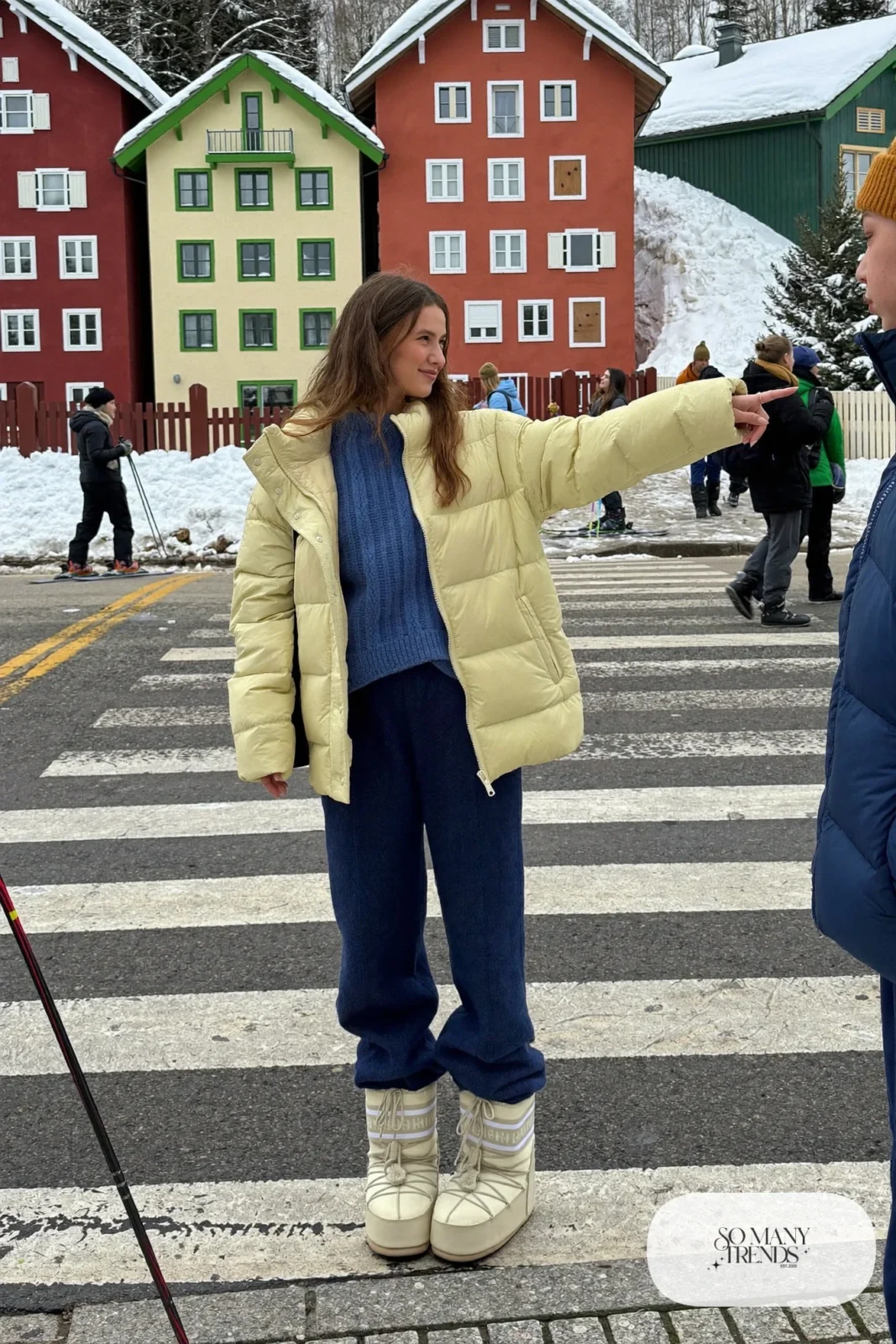 Woman in butter yellow puffer, indigo knit sweater, indigo pants and cream moon boots with a cream beanie in the snow, outfit 25