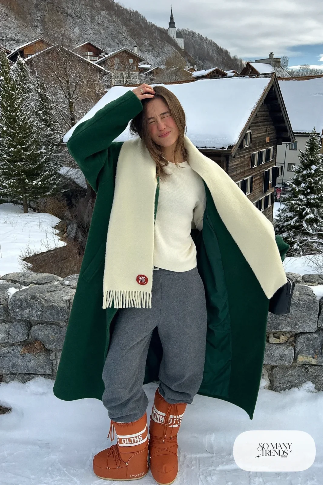 Woman in forest green wool coat, cream sweater, gray pants and terracotta moon boots with a cream scarf in the snow, outfit 27