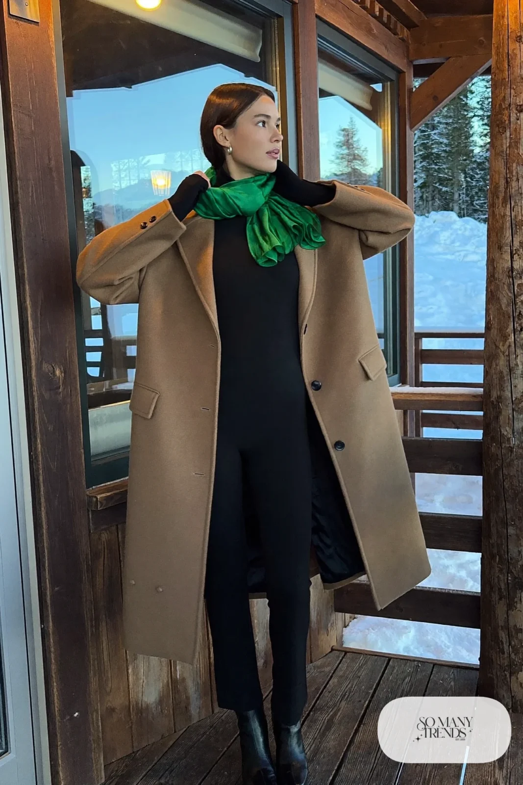 Woman in a black long-sleeve jumpsuit with a belted waist, an oversized camel wool coat, an emerald silk scarf and black ankle boots outside a lodge, outfit 6