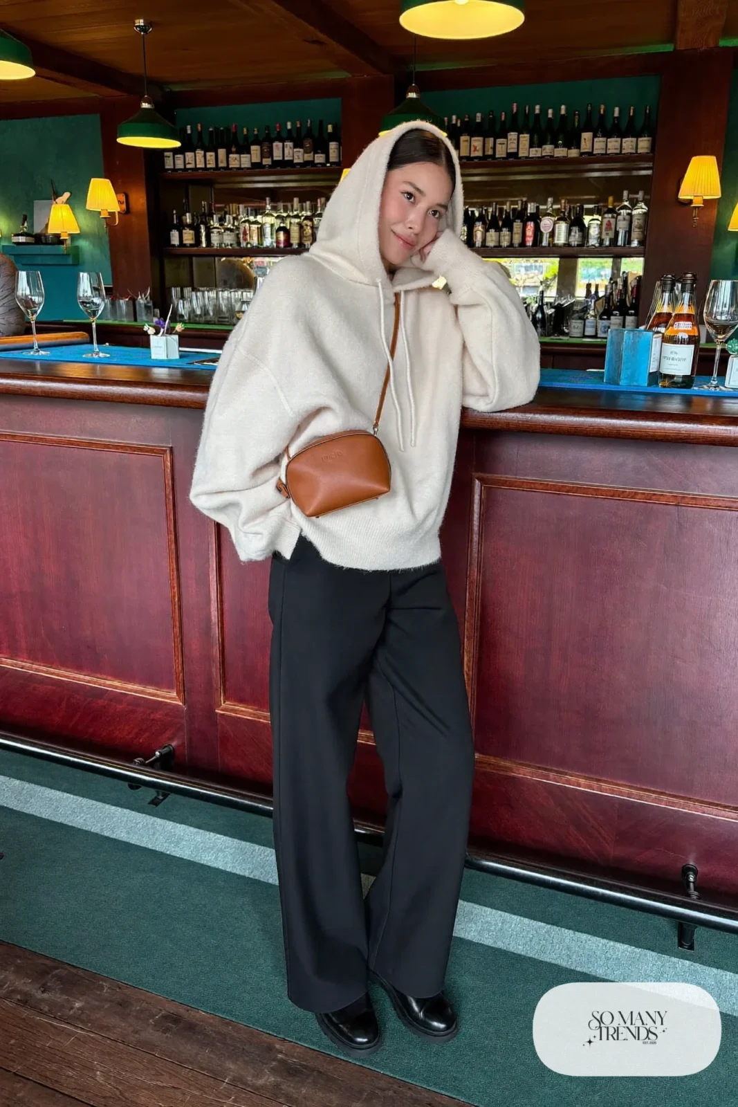 Woman in a pearl cashmere hoodie, black wide-leg wool pants, black lug-sole boots and a cognac belt bag in a lodge bar, outfit 12