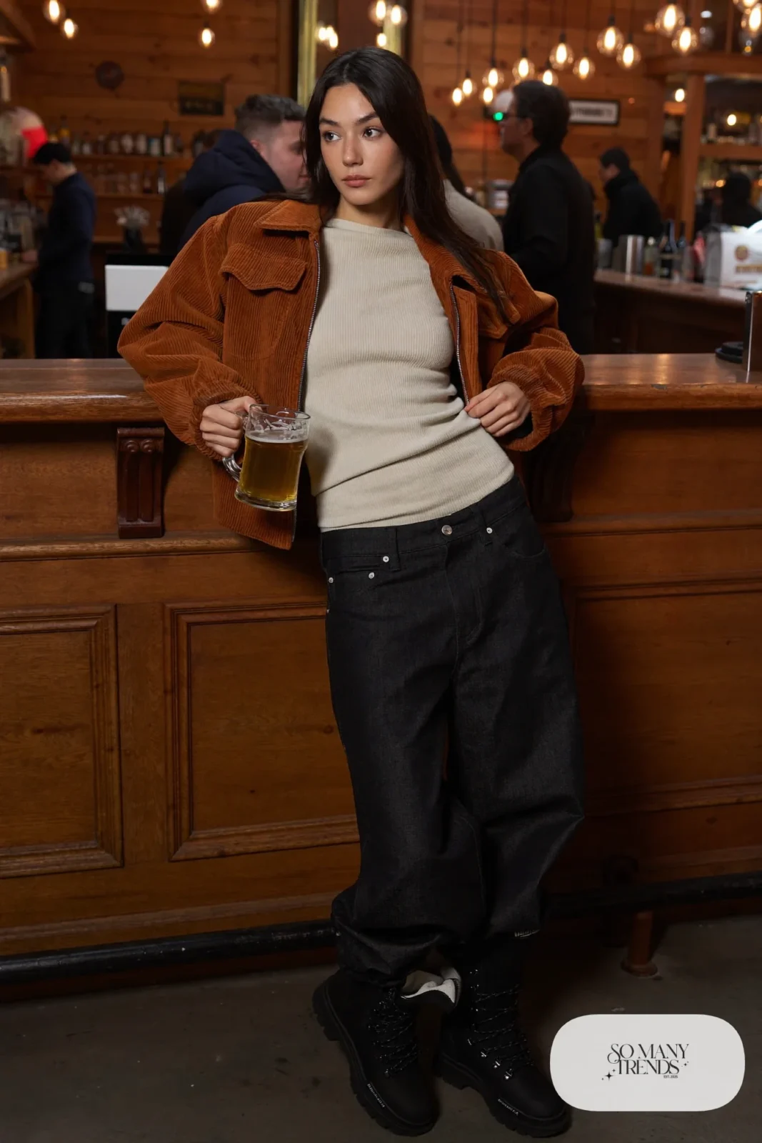 Woman in a cognac corduroy jacket over a cream ribbed sweater with wide black jeans and black lug-sole boots at a lodge bar, outfit 22