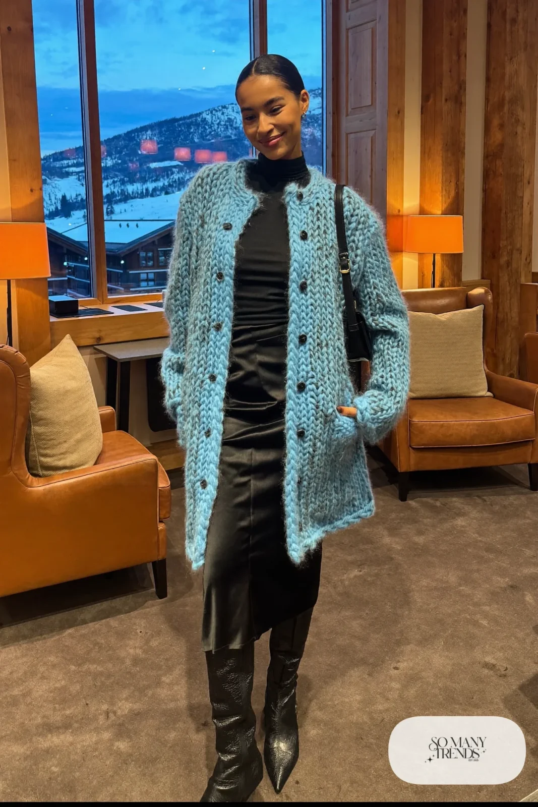 Woman in a long icy blue knit cardigan coat over a black turtleneck and black satin midi skirt with tall black boots in a lodge lounge, outfit 23