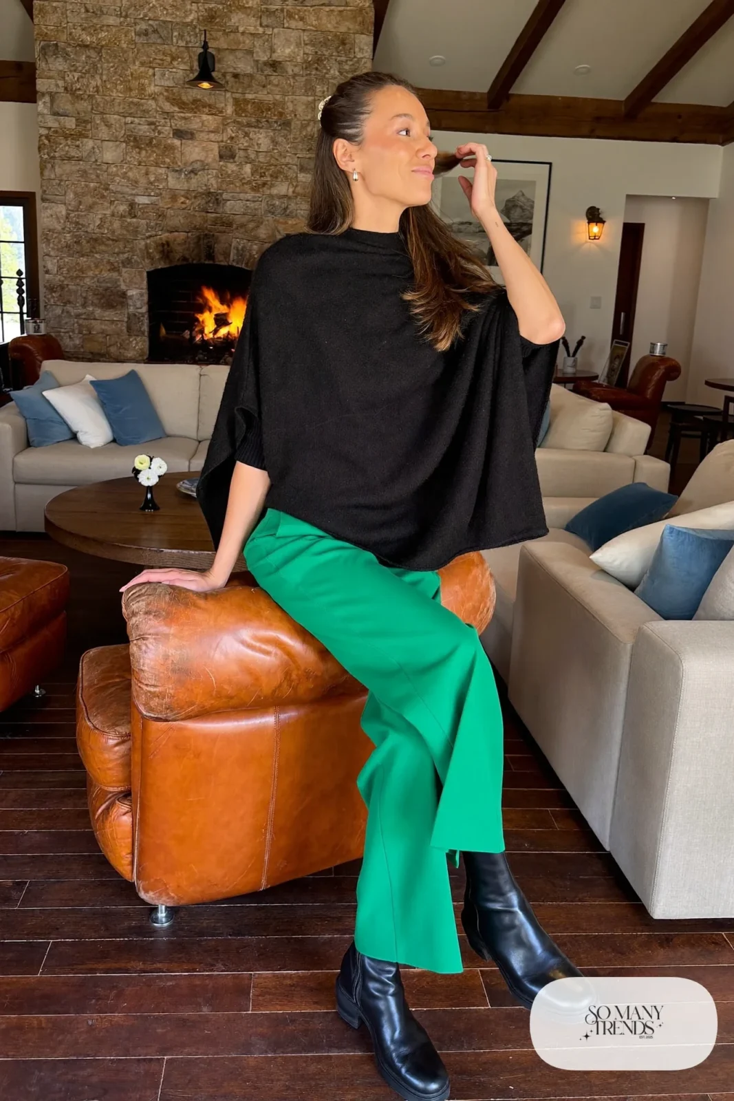Woman in a black cashmere poncho over a fitted black knit top with emerald wide-leg pants and black boots in a lodge lounge, outfit 26