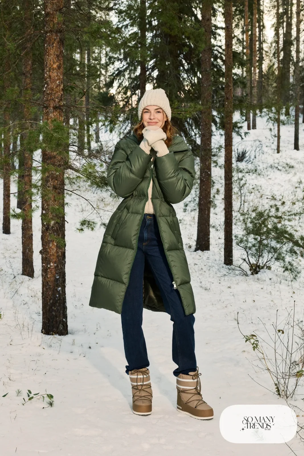 Woman in a long forest green puffer coat, cream beanie, cream mittens, dark jeans and snow boots in a pine forest, outfit 4