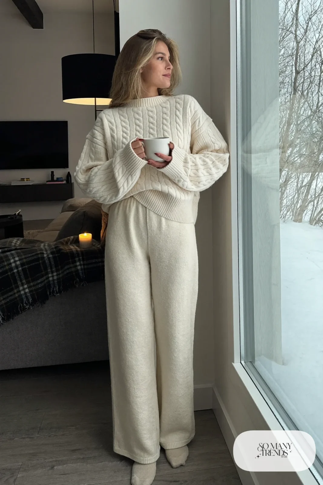 Woman in a cream cable knit sweater and matching straight knit pants with wool socks by a window at home, outfit 7